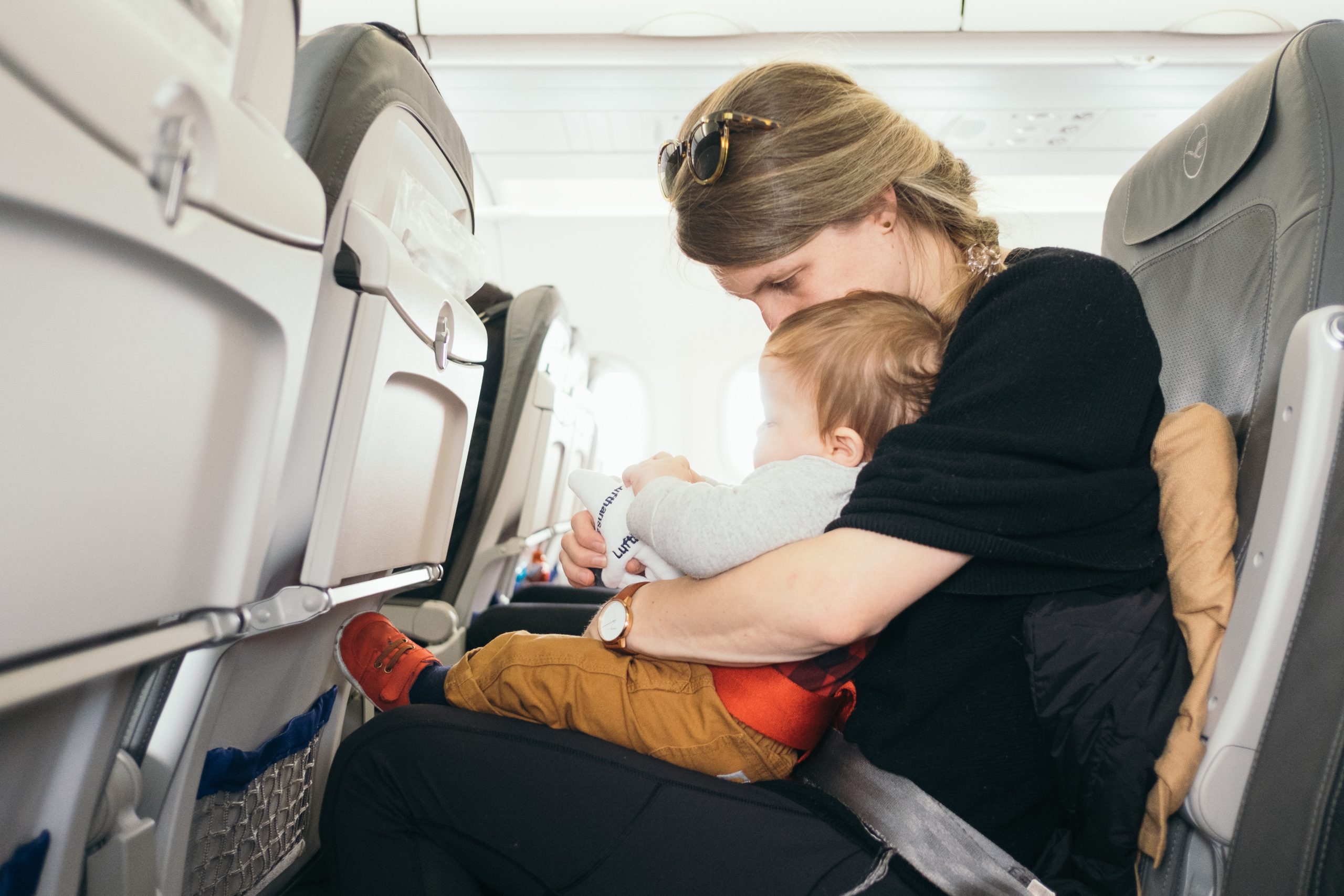 14 Ways to improve your experience at the airport 9 A woman with glasses on her head sits in an airplane seat, holding a baby on her lap. The baby is focused on something in their hands. Both are fastened with a seatbelt. The airplane's interior and rows of seats are visible.
