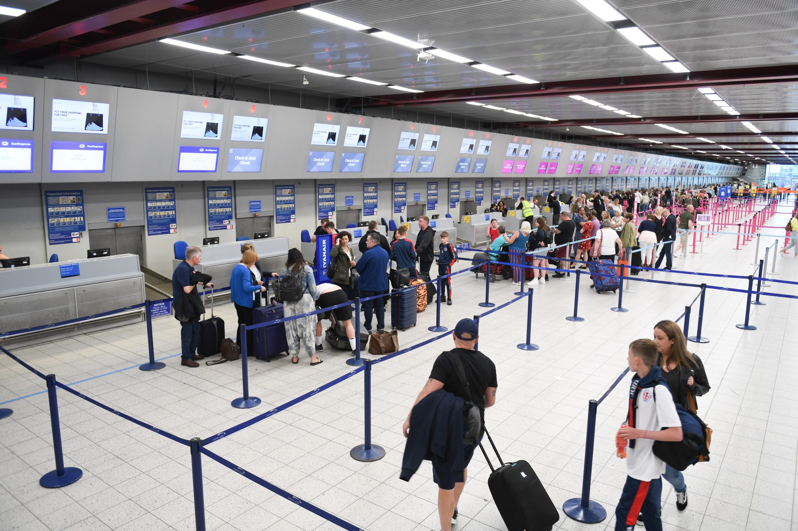 14 Ways to improve your experience at the airport 8 A busy airport check-in area with multiple counters and digital screens. Passengers are standing in line, managing luggage, and waiting to check in. Blue stanchions organize the queues, and staff are assisting travelers. The area is brightly lit.