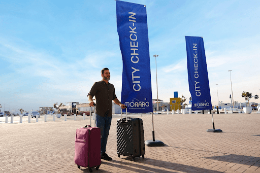 A man stands outside with two rolling suitcases. He is near two large blue banners that read "CITY CHECK-IN". The sky is clear and the ground is paved. There are a few structures and a palm tree visible in the background.